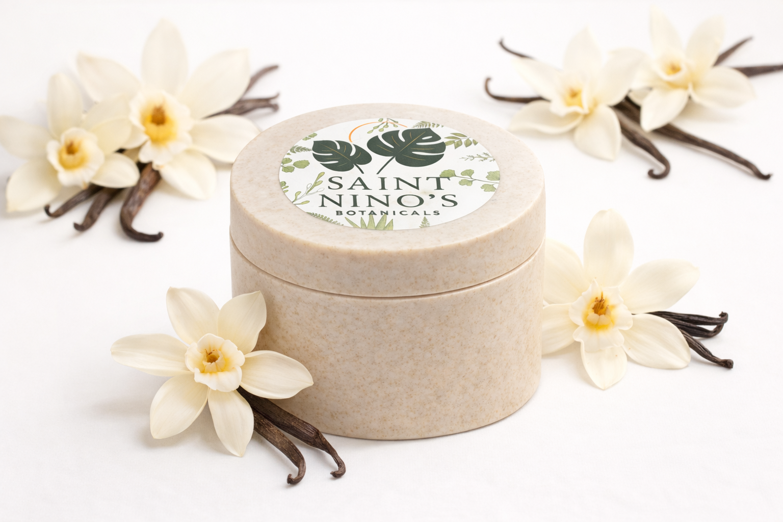 Closed jar of Saint Nino’s Botanicals body butter in a beige container with branded lid, surrounded by vanilla flowers and vanilla pods.